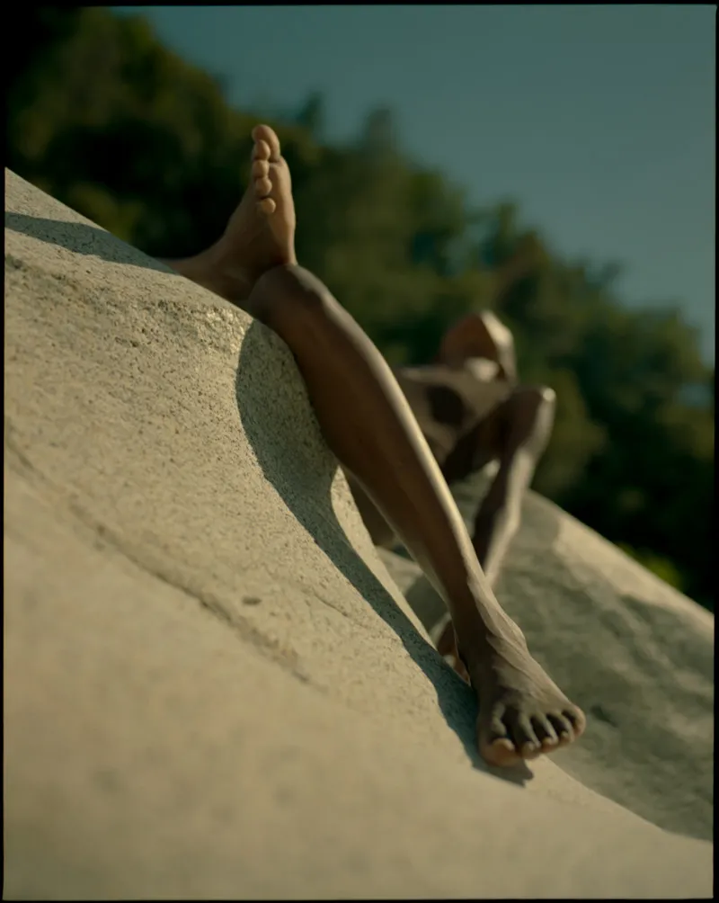 Low angle of the model's bare legs and feet stretched along a sunlit granite slope under blue sky