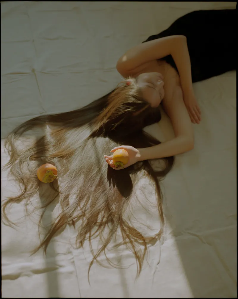 Model in a black dress lying on a white sheet, long hair fanned out, holding a persimmon in sunlight
