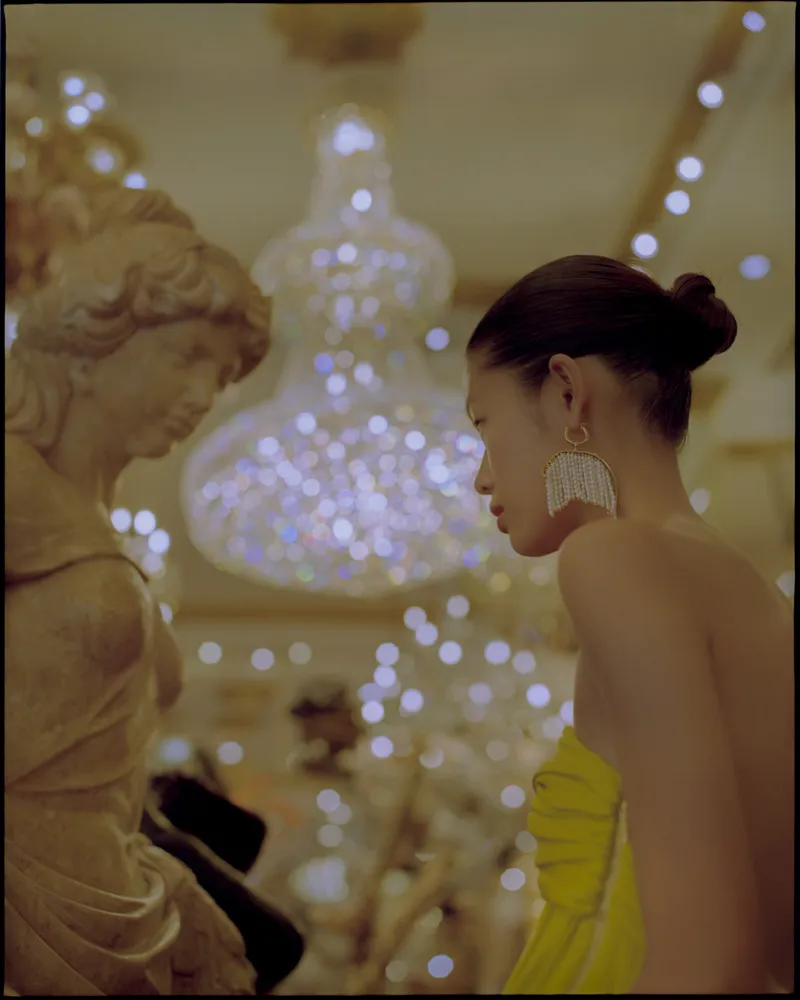 Model in profile in a yellow strapless dress and fringed earring facing a stone statue under a glowing chandelier