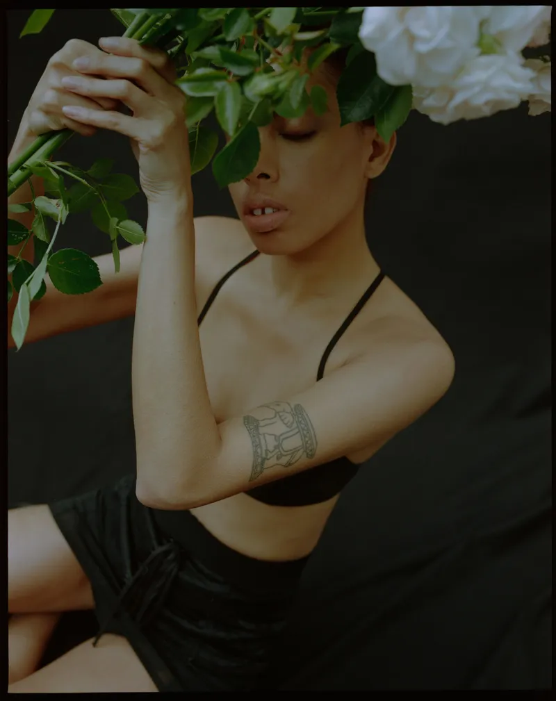 Model in a black bra with an arm tattoo, eyes closed, holding white roses and leaves over the face