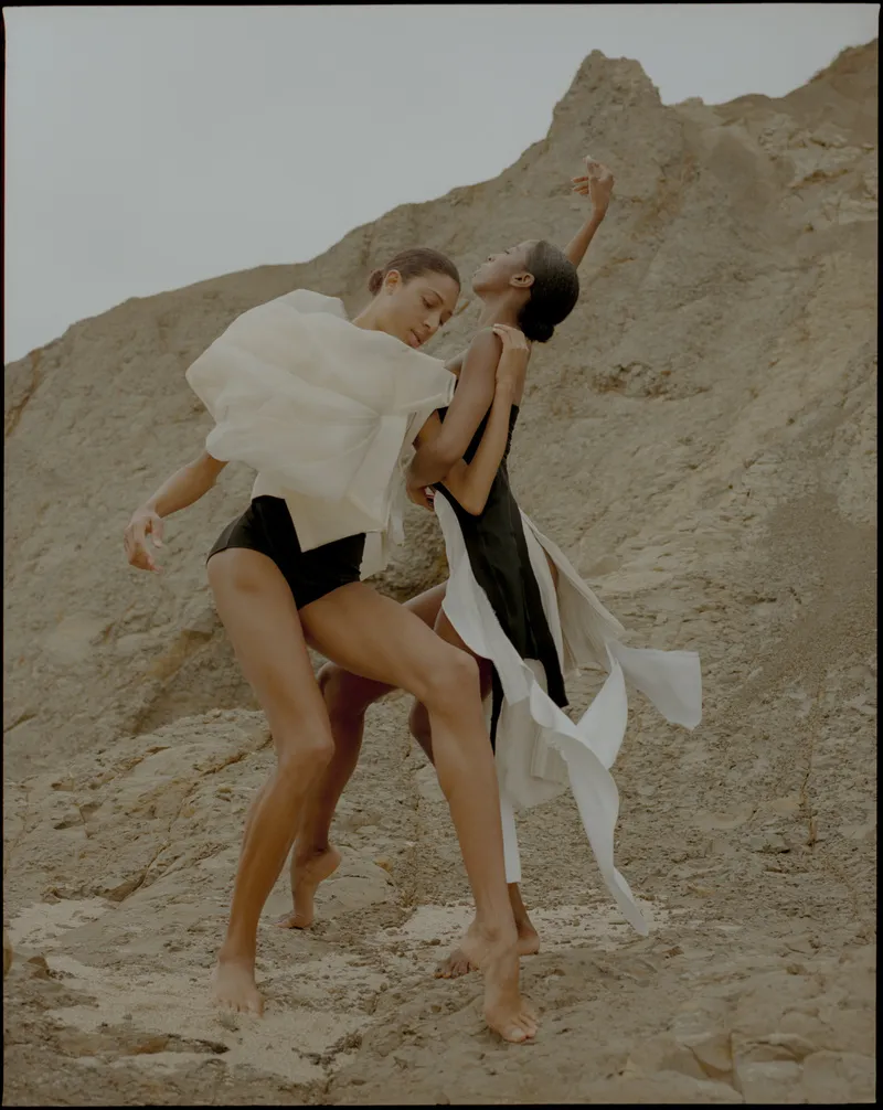 Two models dancing together barefoot on a rocky slope, one in white organza, one in a black and white dress