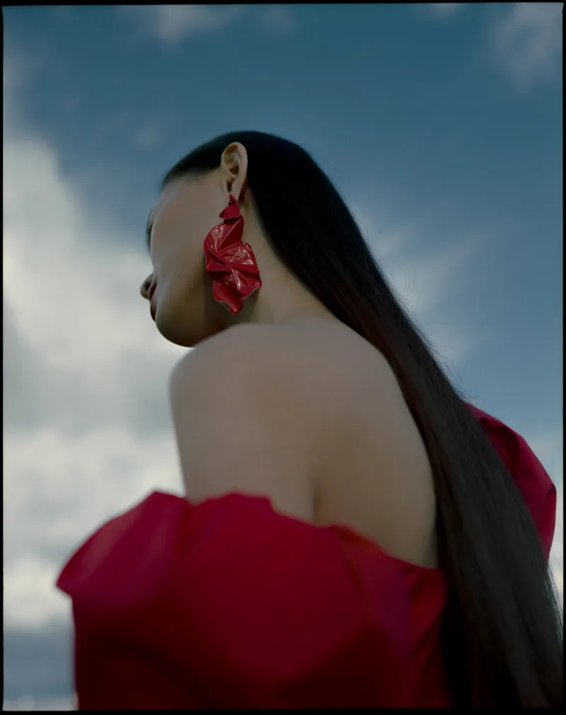 Model from below in an off-shoulder red top with a crumpled red patent earring against cloudy sky