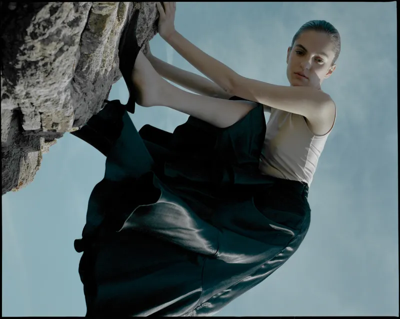 Model in a beige tank and black satin skirt climbing a rock, shot from below against blue sky