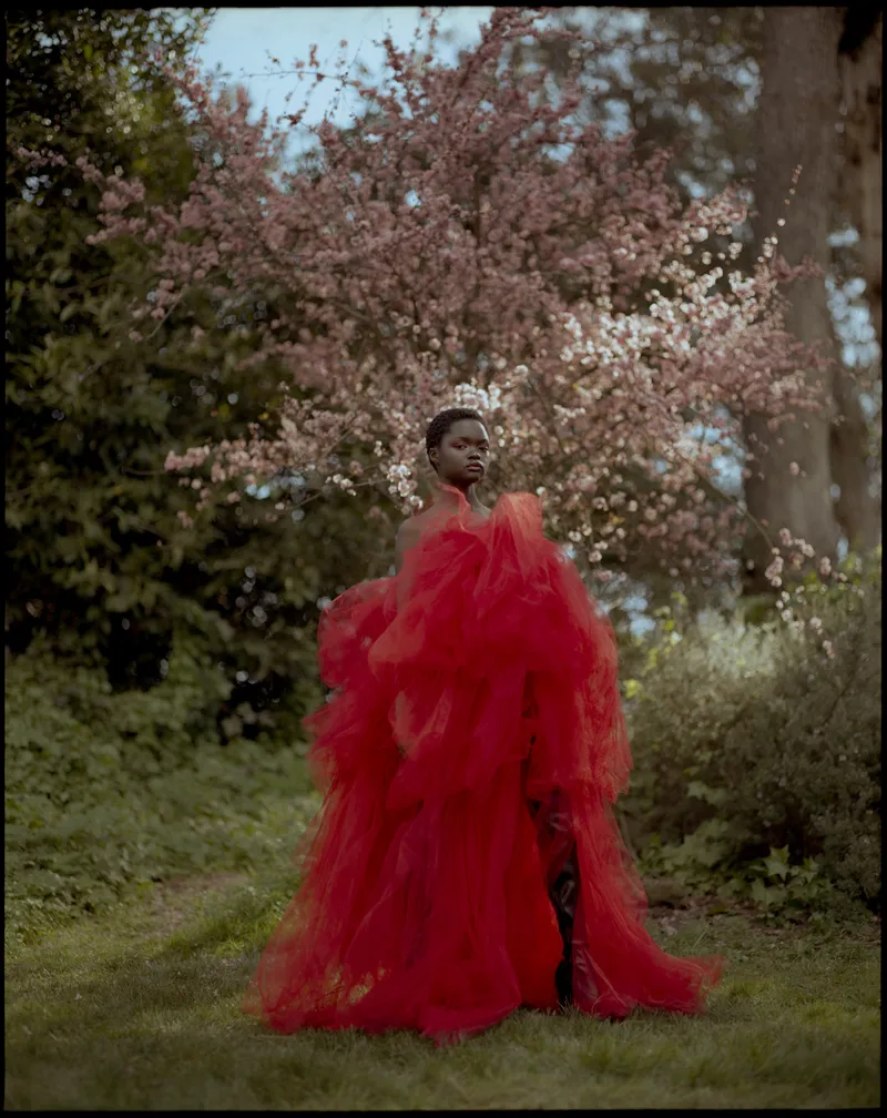 Model in a voluminous red tulle gown standing on grass beneath a pink blossoming tree