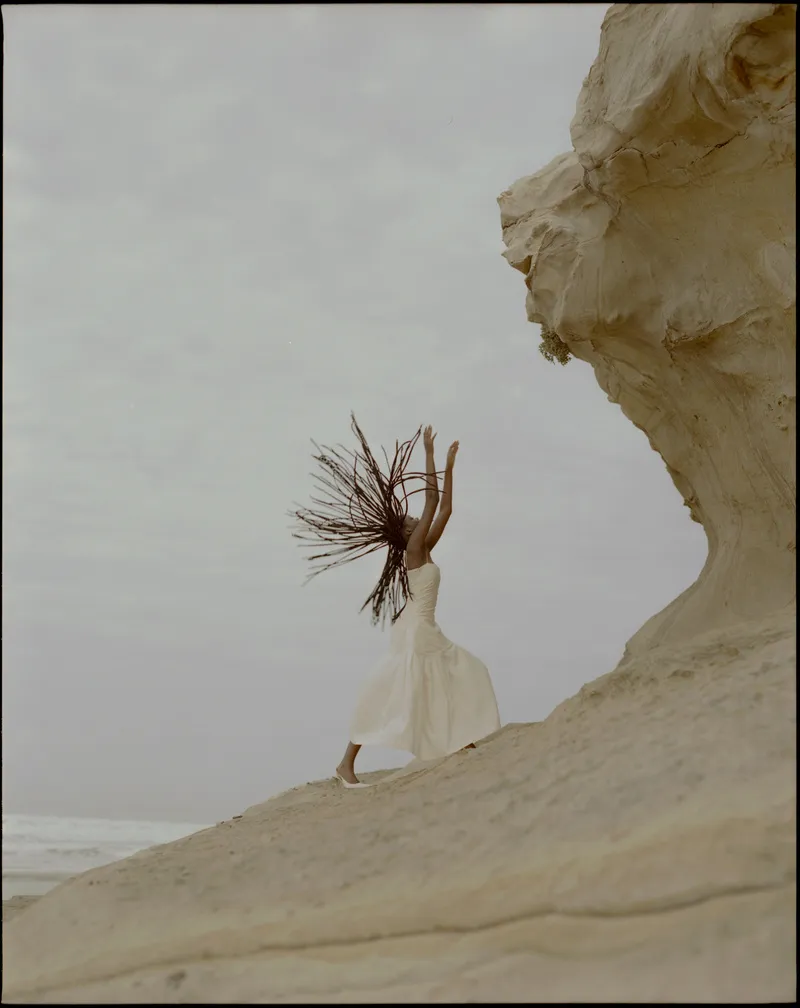 Model in a white drop-waist dress flinging long braids back, arms raised, on a sandstone cliff