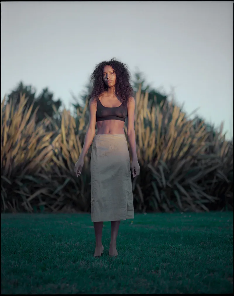 Model in a sheer brown bralette and khaki midi skirt standing barefoot on a lawn before tall flax plants