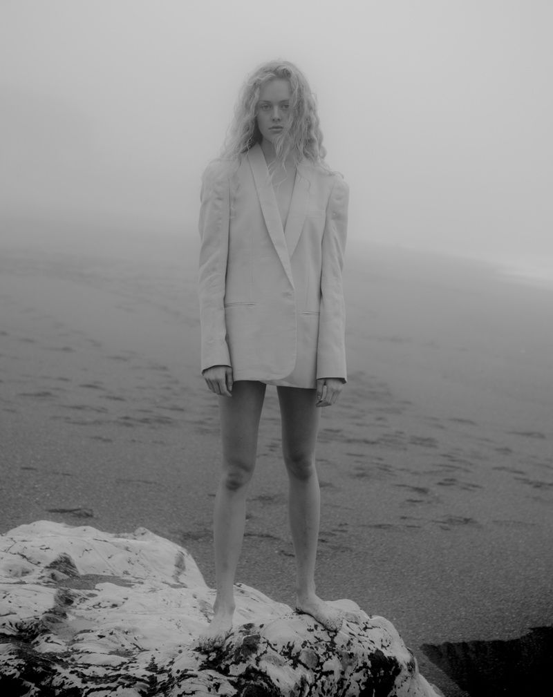 Model in an oversized pale blazer standing barefoot on a white rock on a foggy beach, in black and white