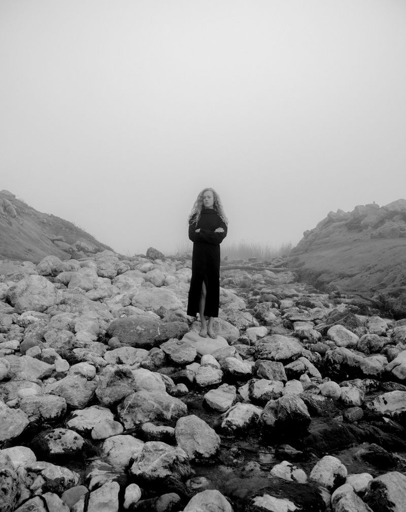 Model in a long black slit dress, arms crossed, standing on a boulder field in fog, black and white
