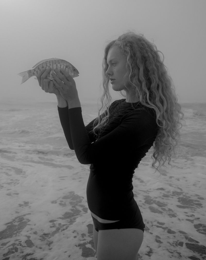 Model in a black long-sleeve swimsuit holding a small fish up in both hands at the foggy shoreline