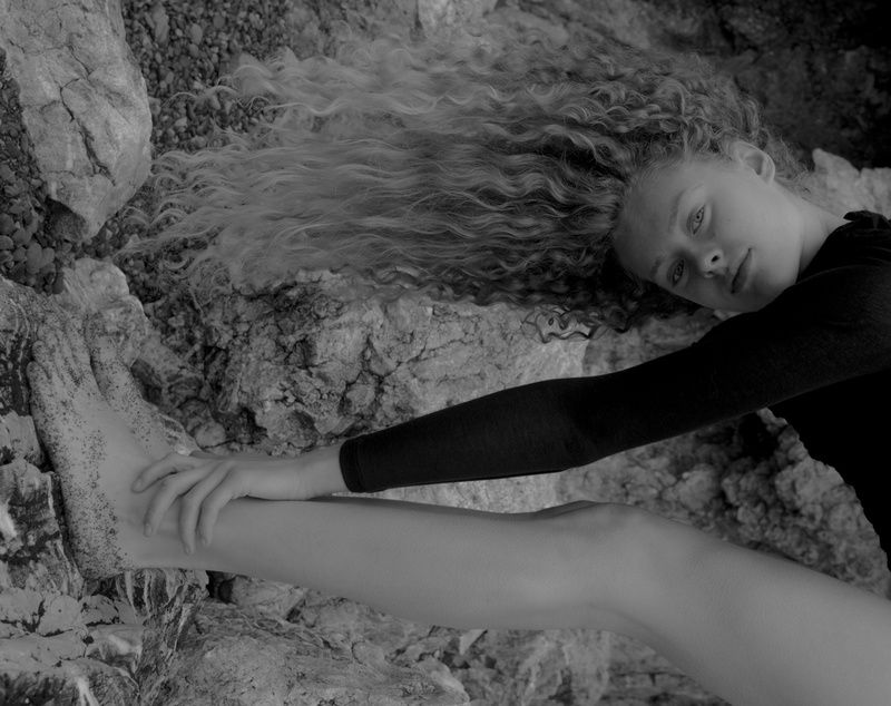 Model reclining on rocks with long curly hair fanned out, hand on an outstretched leg, in black and white