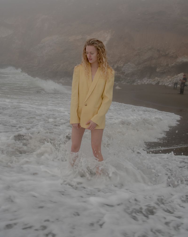 Model in an oversized pale yellow blazer standing knee-deep in foaming surf on a misty beach