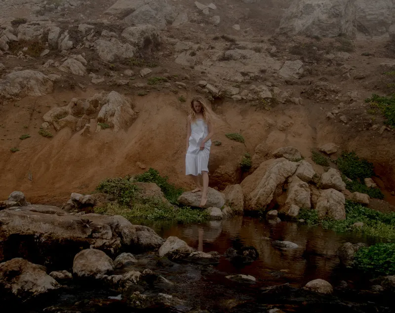 Model in a white slip dress stepping down a sandy rock bank toward a dark tide pool